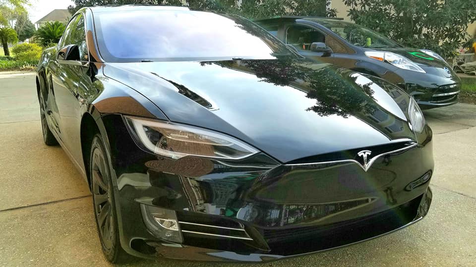 A black Tesla so shiny you can see the sky like a mirror from the paint being polished and ceramic coated to protect for 1 year by our mobile detail vehicle, shown in the background.